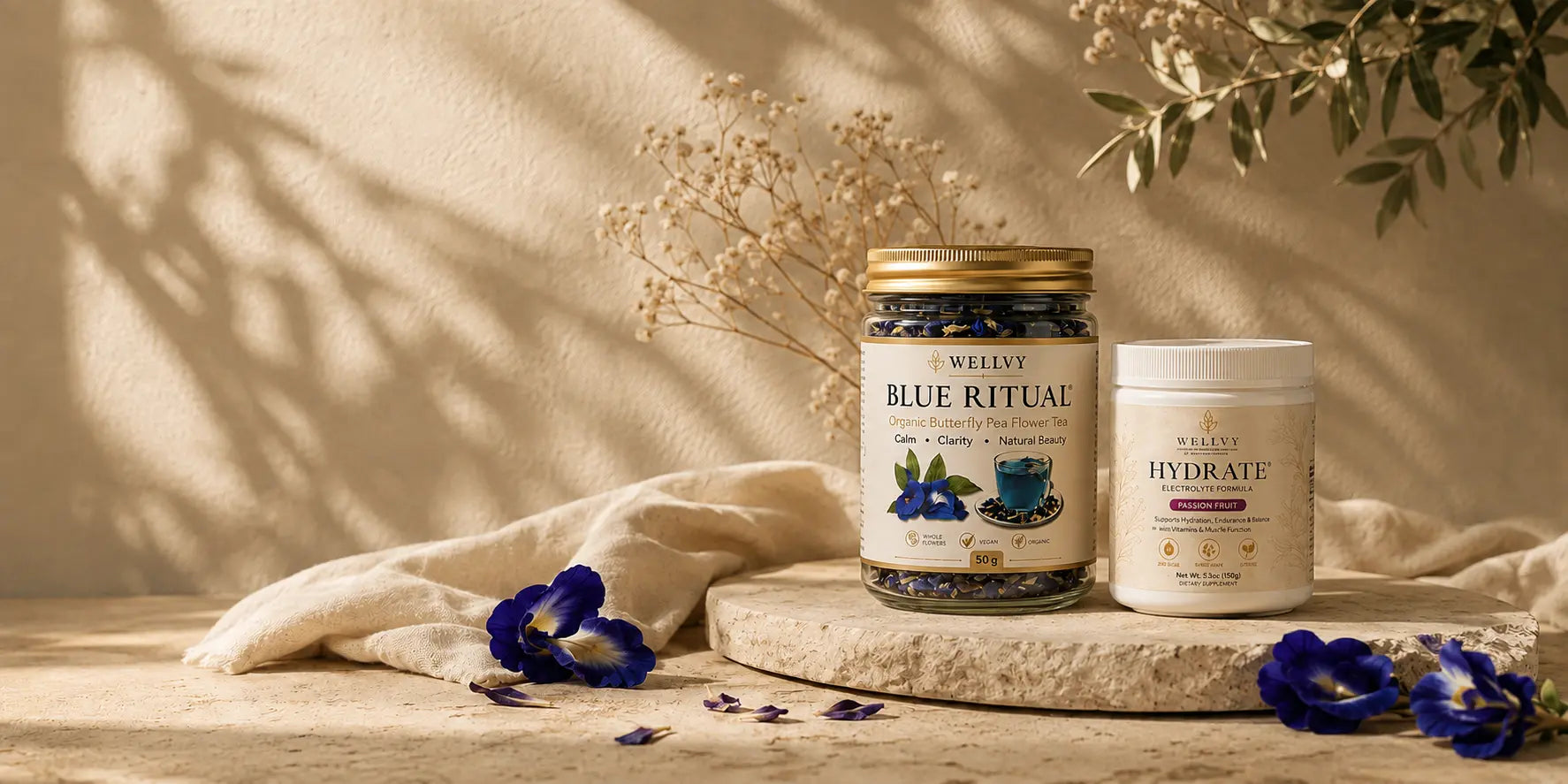 Two wellness products displayed on a stone slab: a glass jar labeled Wellvy Blue Ritual with a gold lid and a white tub labeled Acqua Hydrate, styled with beige fabric, dried flowers, and blue flowers against a warm beige wall with leaf shadows.